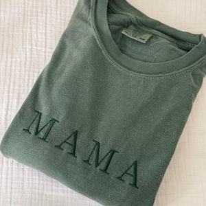 May include: A folded, olive green t-shirt with the embroidered word "MAMA" in a darker green thread. The shirt is made of a soft material and is displayed on a white, textured surface. The text "PICTURED WITH HUNTER THREAD" is in the bottom right corner.