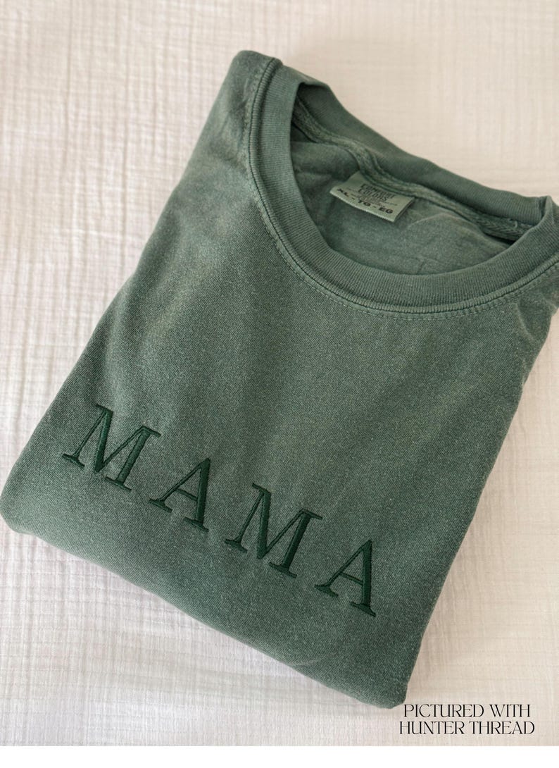 May include: A folded, olive green t-shirt with the embroidered word "MAMA" in a darker green thread. The shirt is made of a soft material and is displayed on a white, textured surface. The text "PICTURED WITH HUNTER THREAD" is in the bottom right corner.