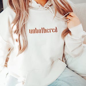 Op de afbeelding: Cr&egrave;mekleurige hoodie met het woord "unbothered" geborduurd in een roodbruin gotisch lettertype over de borst. De hoodie heeft een capuchon met trekkoord en een voorzak. De persoon draagt een lichtblauwe jeans.