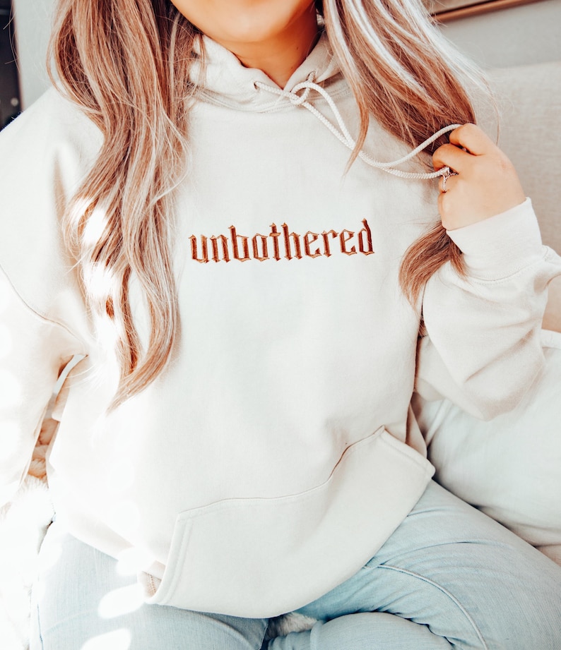 Op de afbeelding: Cr&egrave;mekleurige hoodie met het woord "unbothered" geborduurd in een roodbruin gotisch lettertype over de borst. De hoodie heeft een capuchon met trekkoord en een voorzak. De persoon draagt een lichtblauwe jeans.