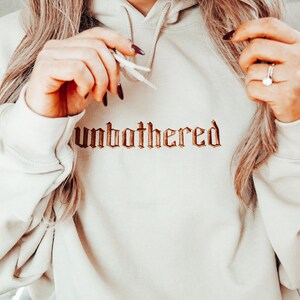 Op de afbeelding: Een cr&egrave;mekleurige hoodie met het woord "unbothered" geborduurd in een bruin, gotisch lettertype over de borst. De hoodie heeft een capuchon met trekkoord en lange mouwen. De afbeelding toont een close-up van de hoodie.