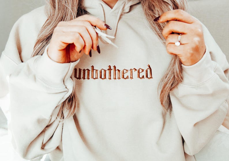 Op de afbeelding: Een cr&egrave;mekleurige hoodie met het woord "unbothered" geborduurd in een bruin, gotisch lettertype over de borst. De hoodie heeft een capuchon met trekkoord en lange mouwen. De afbeelding toont een close-up van de hoodie.