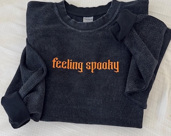 Puede incluir: Camiseta negra de manga larga acanalada con la frase "feeling spooky" en letras naranjas. La camiseta tiene cuello redondo y puños doblados. La tela tiene un aspecto texturizado y vintage.