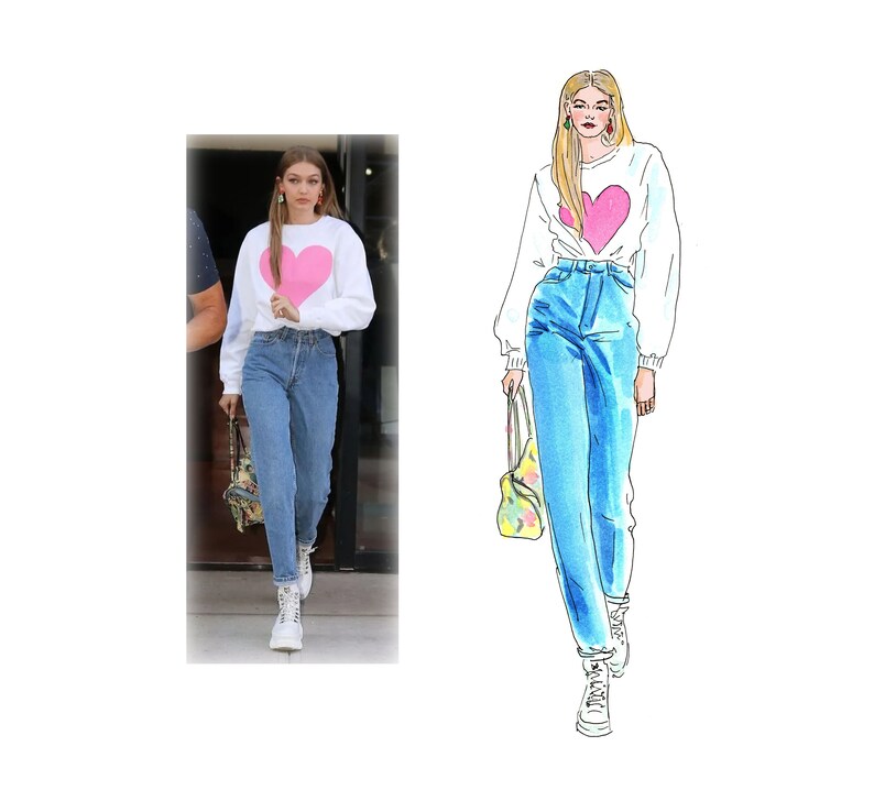 Op de afbeelding: Een vrouw draagt een witte trui met een roze hartontwerp, blauwe jeans en witte sneakers. Ze draagt een tote bag met een bloemenpatroon.