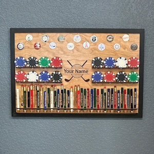 May include: Wooden golf-themed display case with a black frame. It features shelves for golf pencils, poker chips in various colors, and golf ball markers. The text "Your Name" is displayed with crossed golf clubs.