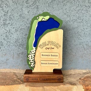 May include: A wooden golf-themed trophy featuring a course layout with green and blue accents. The trophy includes the text "WOLFPACK Golf Club" and the names "Rodney Baker" and "Bryan Sonderby." The base reads "2025 CHAMPIONS."