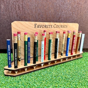 May include: A wooden stand with the words "FAVORITE COURSES" engraved on it. The stand holds 18 pencils, each with a different golf course logo printed on it.