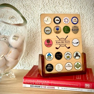 Pode incluir: Um suporte de madeira para bolas de golfe com uma variedade de marcadores de bolas de golfe. O suporte está em cima de uma pilha de três livros. O livro superior é vermelho e tem o título "Harvey Penick's Little Red Book". O livro do meio é vermelho e tem o título "Harvey Penick with Bud Shrake". O livro inferior é vermelho e tem o título "Ben Hogan's Five Lessons". Os marcadores de bolas de golfe têm vários desenhos, incluindo um taco de golfe, uma bola de golfe, um campo de golfe e uma bandeira de golfe. O suporte tem as palavras "Kiss My Putt" impressas nele.