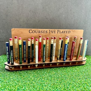 May include: A wooden stand with the text "COURSES I'VE PLAYED" engraved on it. The stand holds 20 golf pencil toppers with different golf course names printed on them.