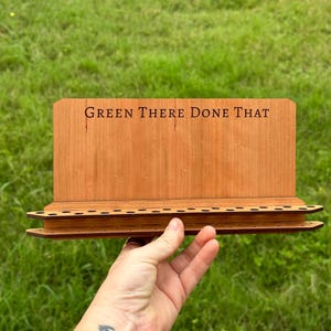 May include: A wooden stand with a sign that reads "Green There Done That" and a row of holes for holding items. The stand is made of light brown wood and has a dark brown stain.
