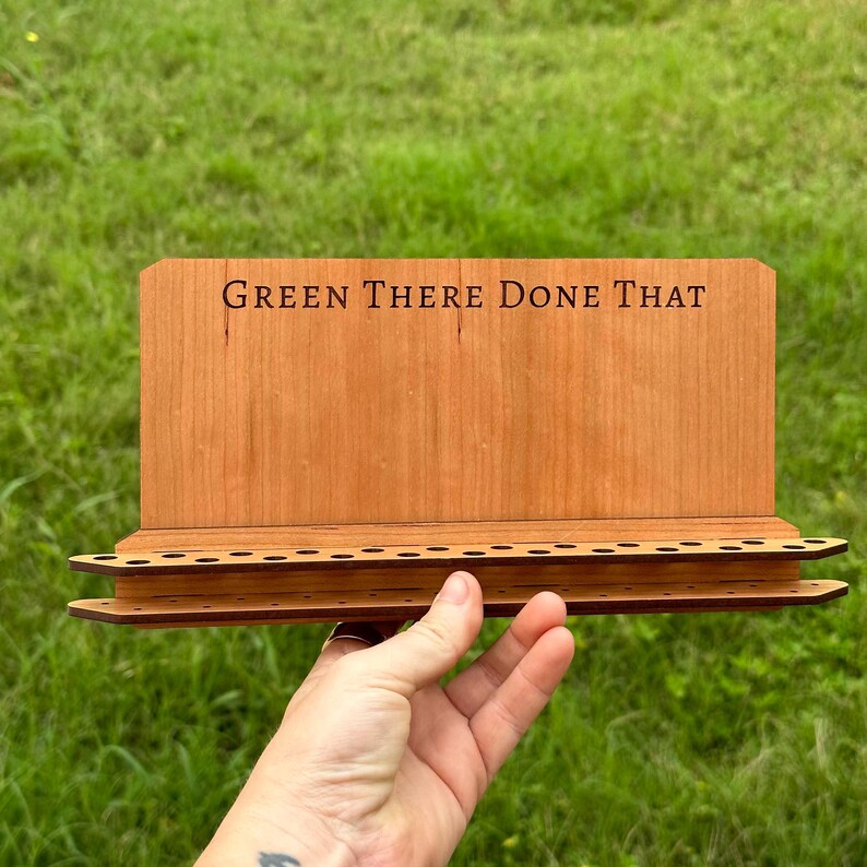 May include: A wooden stand with a sign that reads "Green There Done That" and a row of holes for holding items. The stand is made of light brown wood and has a dark brown stain.