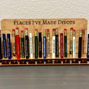 May include: A wooden pencil holder with the text "Places I've Made Divots" on the top. The holder is filled with pencils, each with a different golf course logo and name.
