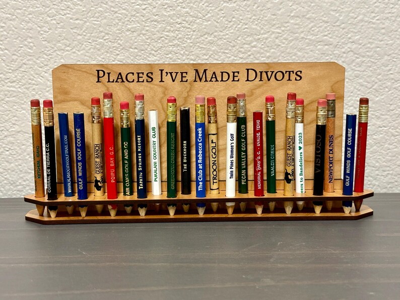 May include: A wooden pencil holder with the text "Places I've Made Divots" on the top. The holder is filled with pencils, each with a different golf course logo and name.