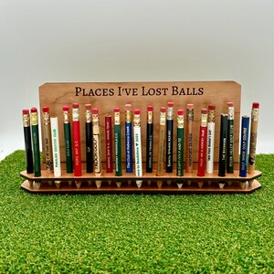 May include: A wooden stand with the text "Places I've Lost Balls" holds a collection of golf pencil toppers with the names of different golf courses printed on them.