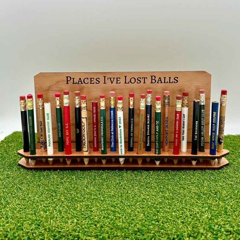 May include: A wooden stand with the text "Places I've Lost Balls" holds a collection of golf pencil toppers with the names of different golf courses printed on them.