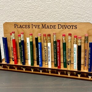 May include: A wooden pencil holder with the text "Places I've Made Divots" on the front. The holder is filled with pencils from various golf courses.
