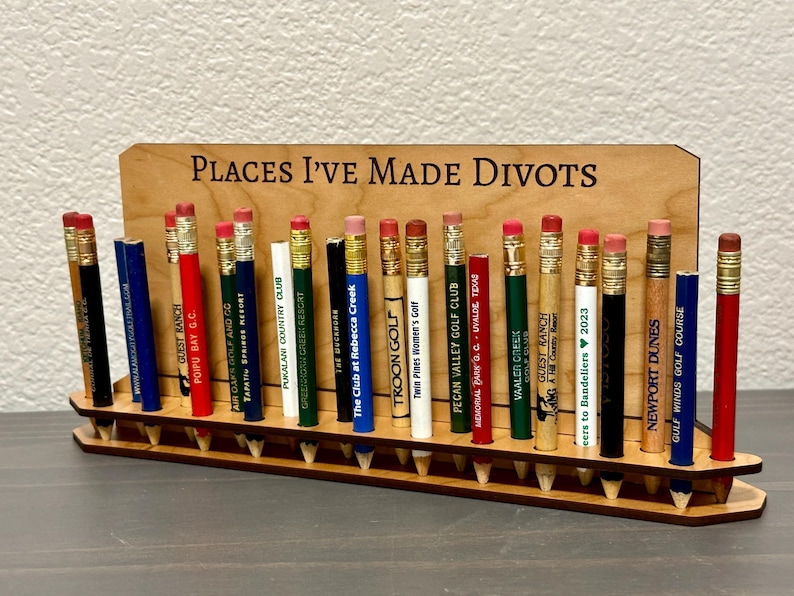 May include: A wooden pencil holder with the text "Places I've Made Divots" on the front. The holder is filled with pencils from various golf courses.