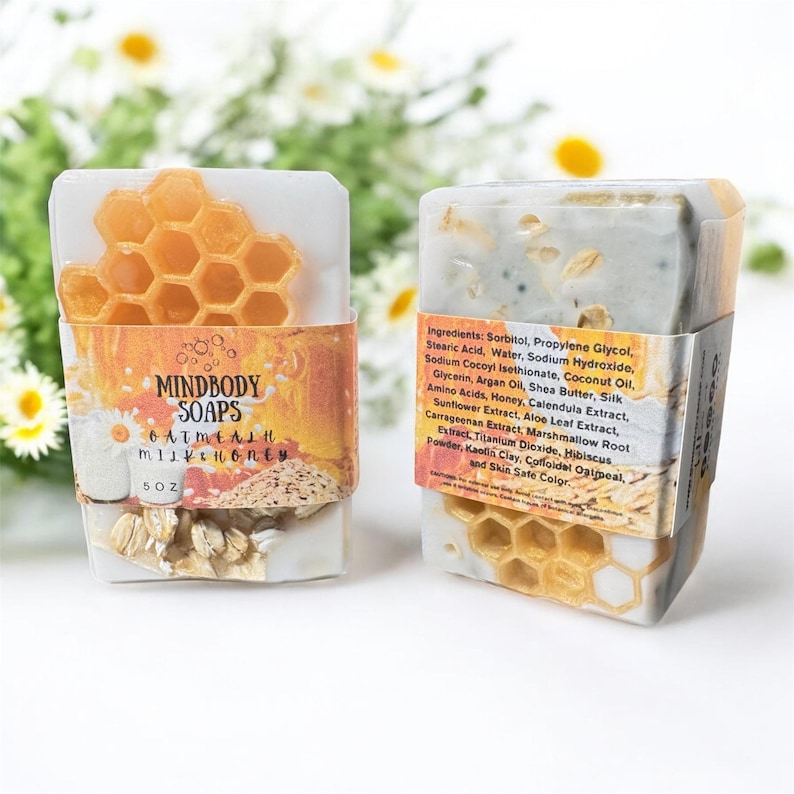 Może przedstawiać: Dwa mydła Mindbody Soaps, Oatmeal Milk & Honey. Mydła w kształcie prostokąta mają wz&oacute;r plastra miodu i etykietę z listą składnik&oacute;w. Mydła są owinięte papierową opaską z nazwą marki i szczeg&oacute;łami produktu.