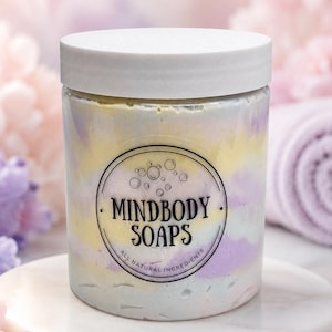 May include: A clear plastic jar filled with swirled pastel yellow, purple, and white cream soap. The jar has a white screw-top lid and a circular label featuring the text MINDBODY SOAPS and ALL NATURAL INGREDIENTS with a bubble graphic.