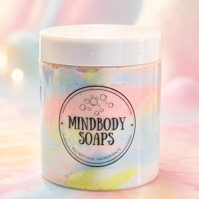 May include: A clear plastic jar with a white lid containing a pastel-colored swirl of soap. The label features a circular design with bubbles and the text MINDBODY SOAPS ALL NATURAL INGREDIENTS in black ink centered on the front of the container.