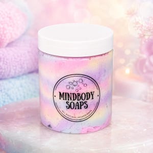 May include: A clear plastic jar filled with pastel-colored whipped soap in swirls of pink, yellow, and blue. The jar has a white screw-top lid and a circular black label featuring the text MINDBODY SOAPS and ALL NATURAL INGREDIENTS.