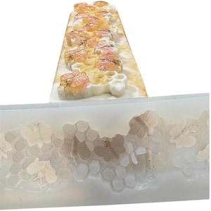 Könnte beinhalten: Eine rechteckige Seifenform mit einem Bienenwaben- und Schmetterlingsdesign. Der obere Teil zeigt Seifenstücke mit rosa und goldenen floralen Akzenten. Der untere Teil zeigt die Form mit detaillierten Schmetterlings- und Bienenwabenmustern.