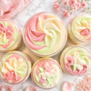May include: Five clear plastic jars filled with whipped body butter, each featuring a swirl of pastel pink, pale yellow, and light green cream. The textures are smooth and piped into peaks, displayed from a top-down perspective.