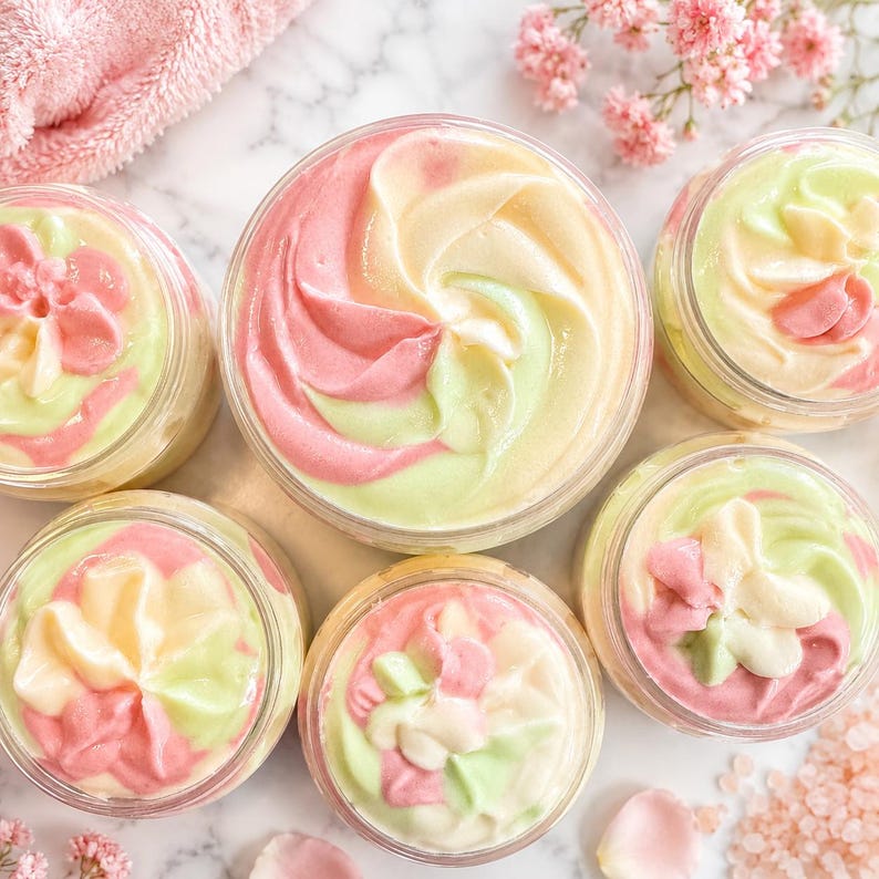 May include: Five clear plastic jars filled with whipped body butter, each featuring a swirl of pastel pink, pale yellow, and light green cream. The textures are smooth and piped into peaks, displayed from a top-down perspective.
