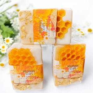 Może przedstawiać: Ułożone w stos kostki mydła z wzorem plastra miodu i napisem "MINDBODY SOAPS OATMEAL MILK & HONEY 5 OZ". Mydło jest jasno-beżowe z ż&oacute;łtym wzorem plastra miodu.