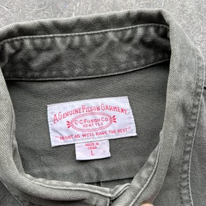 May include: Close-up of a green Filson garment. The label reads "A Genuine Filson Garment" with the text "Might as well have the best." The label also includes the text "C.C. Filson Co. Seattle" and "Made in China L."
