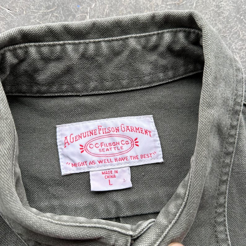 May include: Close-up of a green Filson garment. The label reads "A Genuine Filson Garment" with the text "Might as well have the best." The label also includes the text "C.C. Filson Co. Seattle" and "Made in China L."