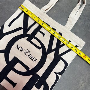 May include: A beige canvas tote bag with black lettering that reads "THE NEW YORKER." The bag is approximately 16 inches wide. The tote has two beige handles and is sitting on a gray surface.