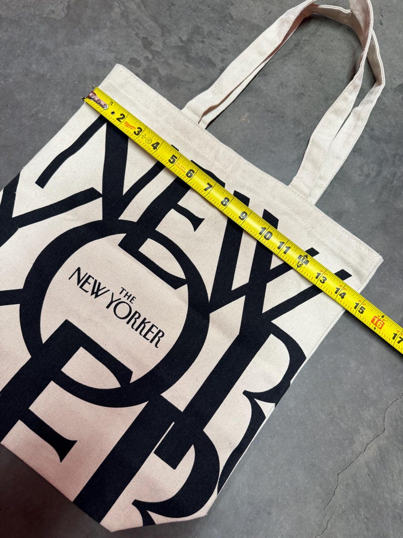 May include: A beige canvas tote bag with black lettering that reads "THE NEW YORKER." The bag is approximately 16 inches wide. The tote has two beige handles and is sitting on a gray surface.