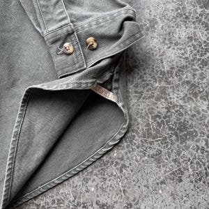 May include: Close-up of a dark green button-down shirt. The shirt features two visible buttons on the cuff and a folded collar. The fabric appears to be a durable cotton or similar material, with visible stitching details.