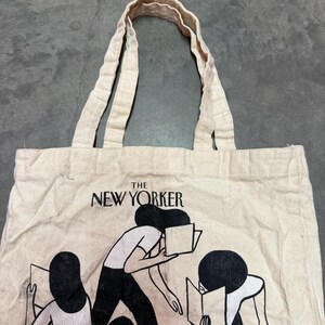 May include: Off-white canvas tote bag featuring the text "THE NEW YORKER" above a graphic of people reading books. The bag has two handles and a simple design, perfect for carrying books, groceries, or everyday essentials.