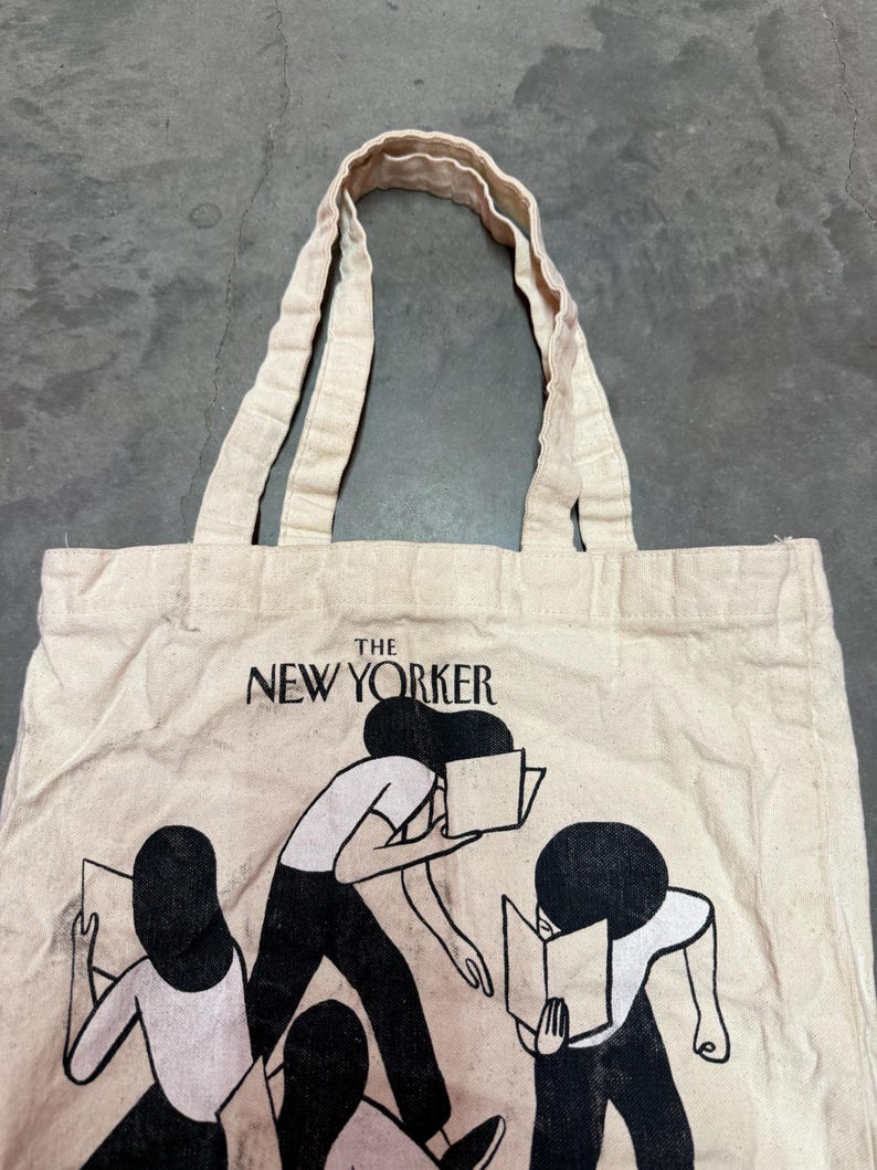 May include: Off-white canvas tote bag featuring the text "THE NEW YORKER" above a graphic of people reading books. The bag has two handles and a simple design, perfect for carrying books, groceries, or everyday essentials.