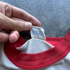 May include: Close-up of a white t-shirt with a red collar and sleeve trim. A tag shows the fabric composition as 50% polyester and 50% cotton, with washing instructions. The shirt is being held by a person.