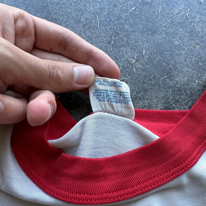 May include: Close-up of a white t-shirt with a red collar and sleeve trim. A tag shows the fabric composition as 50% polyester and 50% cotton, with washing instructions. The shirt is being held by a person.