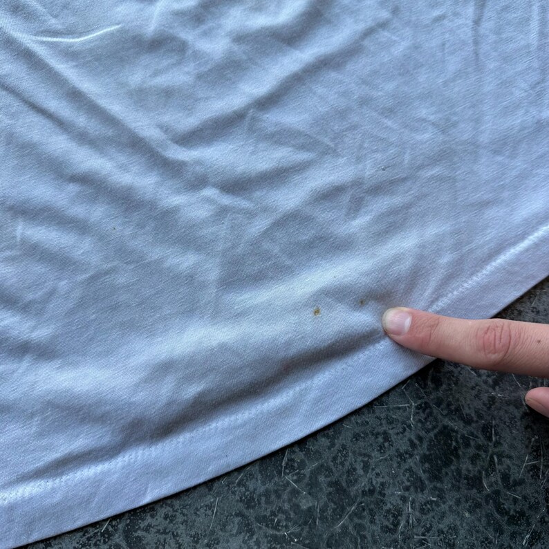 May include: Close-up of a white t-shirt with visible fabric texture and slight discoloration. The bottom hem is visible, with a finger touching the fabric. The shirt appears to be made of a soft material.