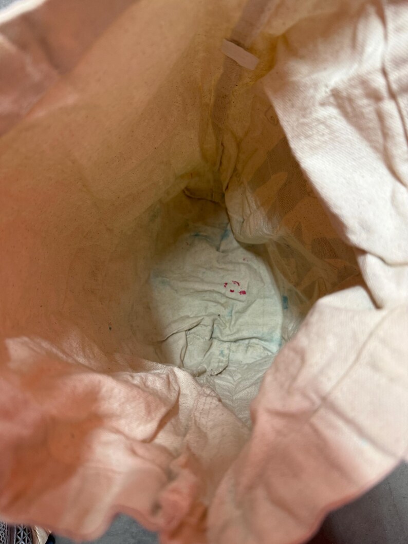 May include: An inside view of a beige canvas bag. The bag's interior is visible, showing the texture of the fabric. The bottom of the bag has some blue and red markings.