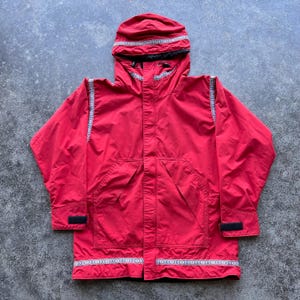 May include: A bright red hooded jacket featuring a black interior lining and a decorative patterned trim in white, blue, and red along the hood, shoulders, and bottom hem. The jacket includes long sleeves with adjustable black velcro cuffs.