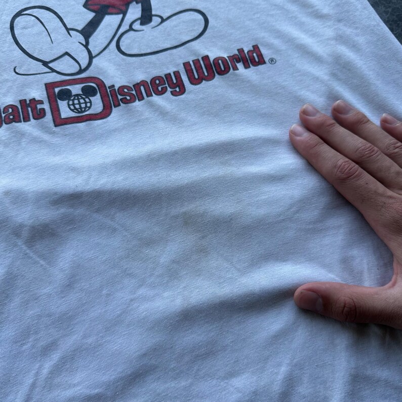 May include: White t-shirt featuring a Mickey Mouse graphic and the text "Walt Disney World" in red. The graphic includes Mickey's feet and the Disney World logo. The shirt is made of a soft, light-colored fabric.