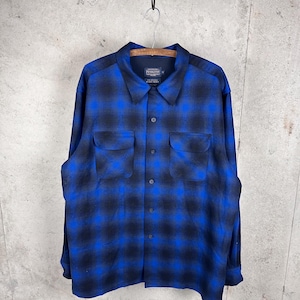 May include: A long-sleeve button-up shirt featuring a blue and black ombre plaid pattern. The shirt includes a pointed collar, a front button placket, and two chest pockets with flaps. The interior label displays the Pendleton brand name.