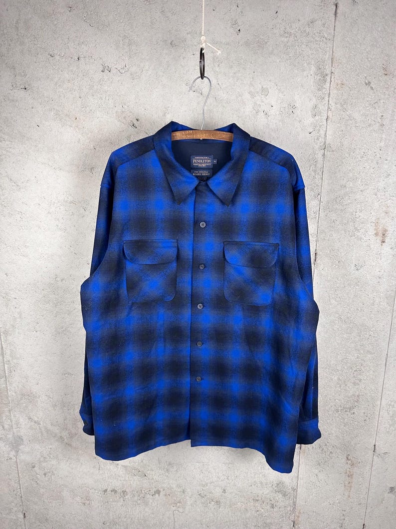 May include: A long-sleeve button-up shirt featuring a blue and black ombre plaid pattern. The shirt includes a pointed collar, a front button placket, and two chest pockets with flaps. The interior label displays the Pendleton brand name.