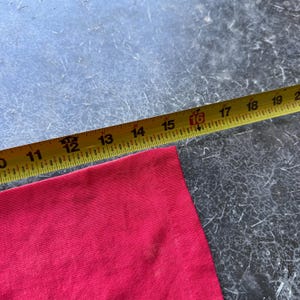 May include: A red fabric corner is measured with a yellow measuring tape. The tape shows measurements from 10 to 20 inches. The fabric is a solid, vibrant red color, and the tape is marked with black numbers.