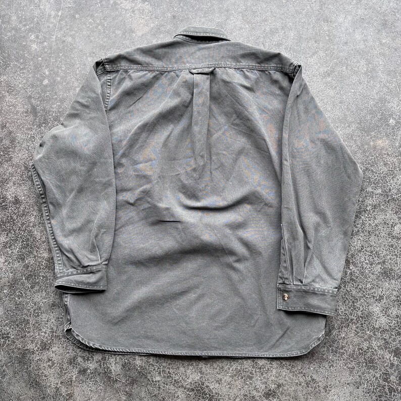 May include: A long-sleeved, button-down shirt in a muted green color. The shirt has a classic collar, buttoned cuffs, and a curved hem. The back of the shirt features a yoke and a box pleat for added comfort and style. The fabric appears to be a soft, washed cotton.