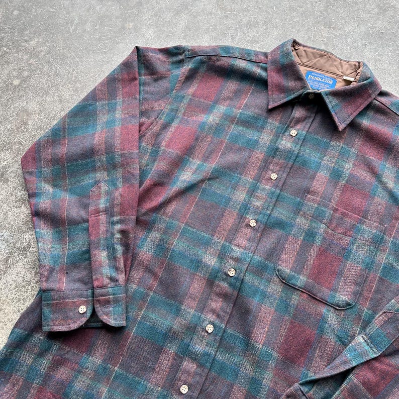 May include: A long-sleeved, button-down shirt with a plaid pattern in shades of teal, burgundy, and gray. The shirt has a collared neckline, a chest pocket, and the brand name "Pendleton" on a tag.