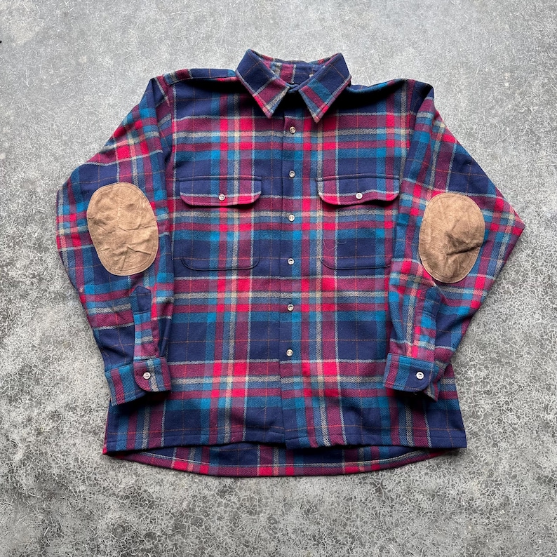K&ouml;nnte beinhalten: Ein lang&auml;rmeliges kariertes Hemd in Marineblau, Rot und T&uuml;rkis, mit zwei Brusttaschen und braunen Ellbogenpatches. Das Hemd hat eine Knopfleiste und Manschetten und ist aus einem weichen, gewebten Stoff gefertigt.