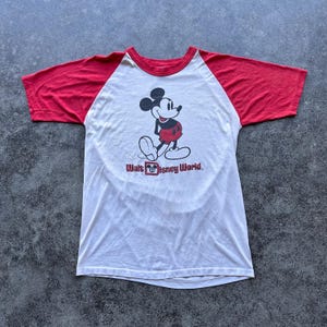 May include: White and red raglan-style t-shirt featuring a graphic of Mickey Mouse with the text "Walt Disney World." The shirt has a red neckline and sleeves, and a white body. The Mickey Mouse graphic is in black, red, and white.