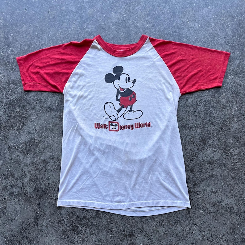 May include: White and red raglan-style t-shirt featuring a graphic of Mickey Mouse with the text "Walt Disney World." The shirt has a red neckline and sleeves, and a white body. The Mickey Mouse graphic is in black, red, and white.